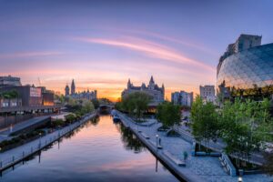 A sunset view of downtown Ottawa featuring the Rideau Canal lined with walkways, trees, and lampposts. The Fairmont Château Laurier and Parliament buildings appear in the background, while the glass façade of the Shaw Centre reflects the colourful sky.